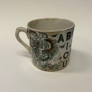 Puede incluir: Taza de cerámica blanca antigua con asa. La taza presenta una ilustración en blanco y negro de una casa, flores y las letras A, B, I, O y U. La taza tiene un aspecto ligeramente envejecido.