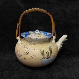 May include: A ceramic teapot with a cream-coloured body featuring painted plum blossom branches and a potted plant. The lid has a blue floral border and a small knob. A curved bamboo handle is attached to the top with metal loops and a spout.