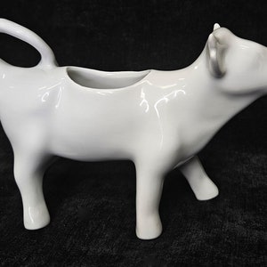 May include: A white ceramic cow-shaped creamer. The creamer features a handle and a spout, and stands on four legs. This cow creamer is a decorative item, designed for serving cream or milk.