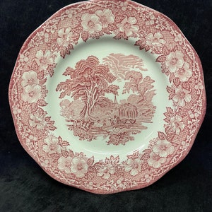 Puede incluir: Un plato blanco y rojo con estampado floral y borde festoneado. El plato presenta una escena central de paisaje en rojo, rodeada por un borde de flores y hojas. El plato es probablemente de cerámica.