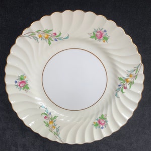 May include: Cream-colored, scalloped-edge plate with a gold rim and a white center. The plate features a spiral design and is decorated with floral sprigs in pink, yellow, blue, and green. A vintage-style decorative plate.