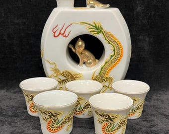 May include: A white ceramic sake set featuring a decorative dragon and cat design. The set includes a bottle with a gold bird on top and six small cups. The dragon design incorporates gold, green, and red accents.