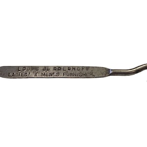 May include: A vintage metal button hook with a pointed end and a curved hook. The text "LOUIS JR. GOLDHOFF LADIES' & MEN'S FURNISHER" is printed on the handle. The button hook is silver in colour.