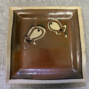 May include: Square ceramic dish with a brown interior and a cream-colored border. The dish features two abstract, cream-colored shapes with black and brown accents. The dish is likely handmade and suitable for serving small portions.