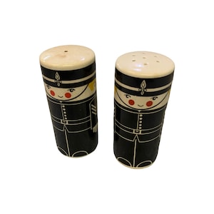 Könnte beinhalten: Ein Paar Vintage-Salz- und Pfefferstreuer. Jeder Shaker ist zylindrisch und hat ein schwarzes Uniformdesign mit einem lächelnden Gesicht, roten Wangen und einem weißen Hut. Die Oberteile sind cremefarben.