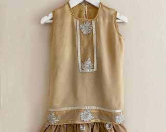 May include: A two-piece outfit for girls in a light brown color with silver embroidery. The top is sleeveless and has a round neckline. The skirt is a-line and has a ruffle at the bottom.