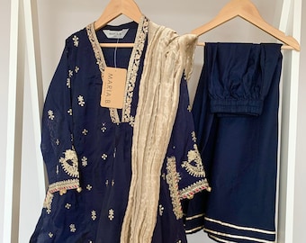 May include: Navy blue embroidered tunic and matching pants with a gold-colored scarf. The tunic features intricate gold and red embroidery along the neckline, sleeves, and hem. The pants have gold trim at the ankles. The outfit is displayed on a wooden hanger.