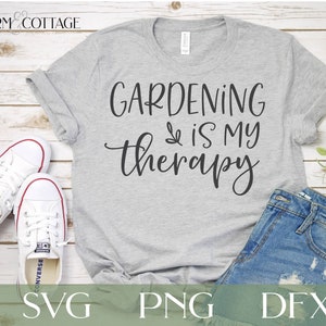 May include: A gray t-shirt with the text "GARDENING is my therapy" in black script font. The shirt is laid flat on a white wood background with a pair of white sneakers and a pair of blue jeans.