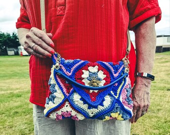 Puede incluir: Una mujer lleva una camisa roja y lleva un bolso de ganchillo con un patrón geométrico azul, blanco y rojo. El bolso tiene una solapa azul con un acento de abeja dorada.