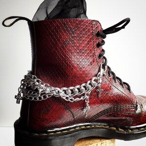Puede incluir: Botas de cuero rojo con detalles de cadena y cruces plateados. Las botas tienen un cierre de cordones negros y una suela gruesa.
