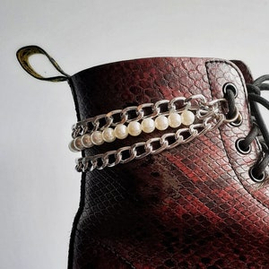 Puede incluir: Un par de botas de cuero rojo oscuro con una cadena plateada y un adorno de perlas alrededor del tobillo.