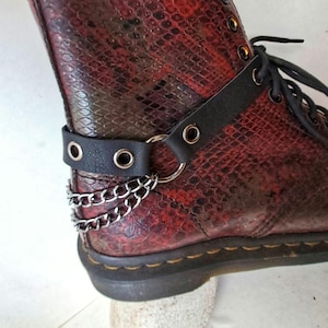 Puede incluir: Primer plano de una bota roja y negra con estampado de piel de serpiente, con una correa de cuero negro y detalles de cadena plateada. La bota tiene cordones negros y una suela negra. La correa se sujeta con ojales plateados.