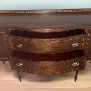 May include: A vintage wooden sideboard with two drawers and two doors. The sideboard has a curved front and is made of a dark wood with a light wood inlay. The sideboard is sitting on a light wood floor.