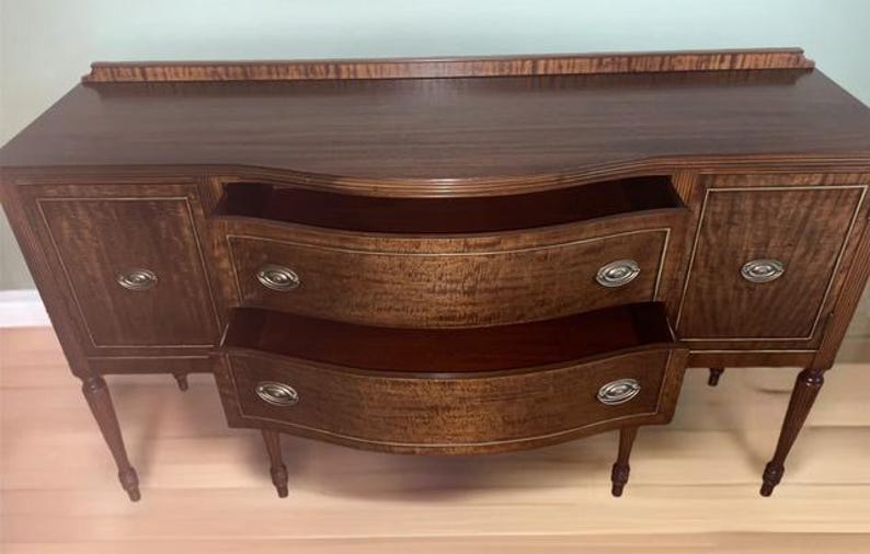 May include: A vintage wooden sideboard with two drawers and two doors. The sideboard has a curved front and is made of a dark wood with a light wood inlay. The sideboard is sitting on a light wood floor.
