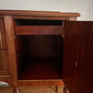 May include: A wooden cabinet with a single drawer and a door that opens to reveal an empty interior. The cabinet has a dark wood finish and decorative trim.