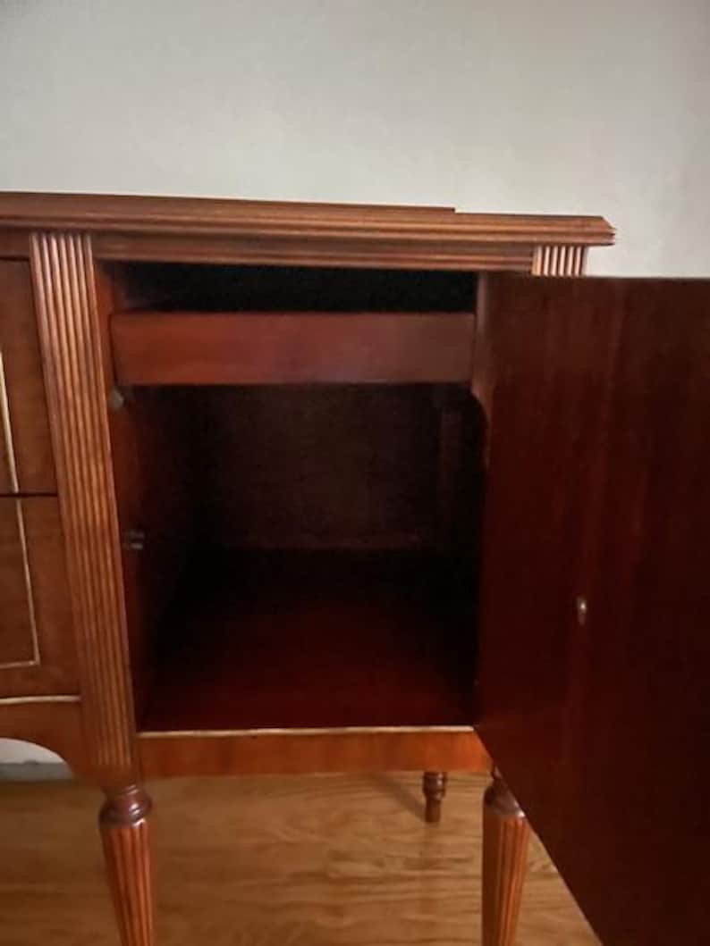 May include: A wooden cabinet with a single drawer and a door that opens to reveal an empty interior. The cabinet has a dark wood finish and decorative trim.