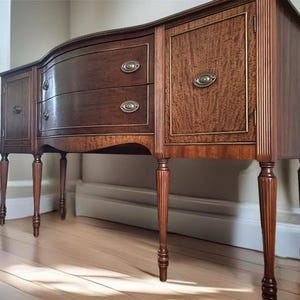 May include: A vintage mahogany wood sideboard with two drawers and three doors. The sideboard has a curved front and sits on four turned legs.