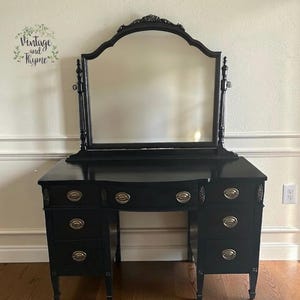 May include: A black vanity table with a matching arched mirror. The piece features five drawers with brass-toned oval pulls, two vertical side supports for the mirror, and a central drawer. The surface is painted in a solid black finish.