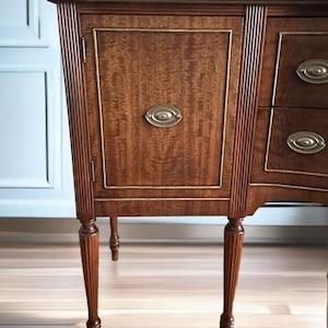 May include: A wooden sideboard with a curved front and fluted legs. The sideboard has a single door on the left side and three drawers on the right side. The door has a brass oval-shaped handle. The sideboard is a rich brown color and has a wood grain pattern.
