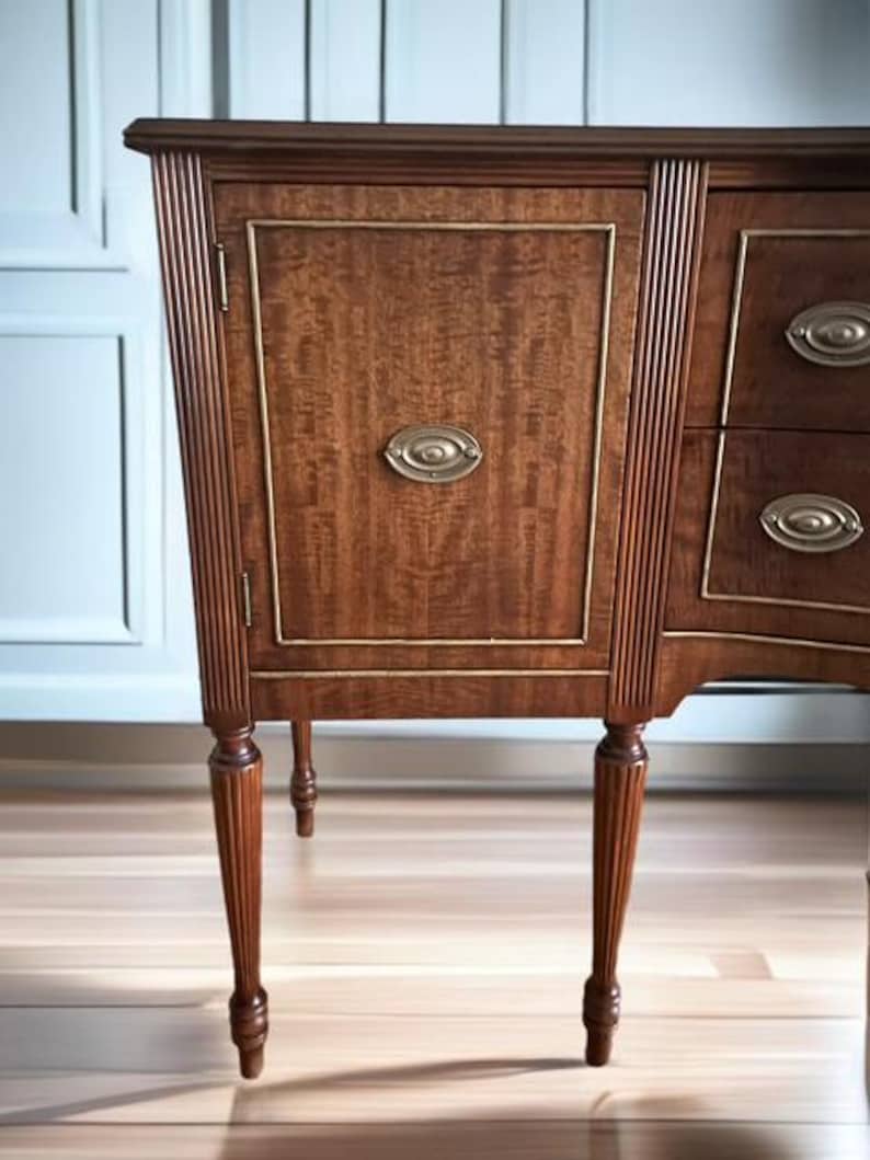 May include: A wooden sideboard with a curved front and fluted legs. The sideboard has a single door on the left side and three drawers on the right side. The door has a brass oval-shaped handle. The sideboard is a rich brown color and has a wood grain pattern.