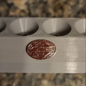 May include: A close-up of a grey plastic mould with six circular indentations. A copper-coloured coin with a design is sitting in one of the indentations.