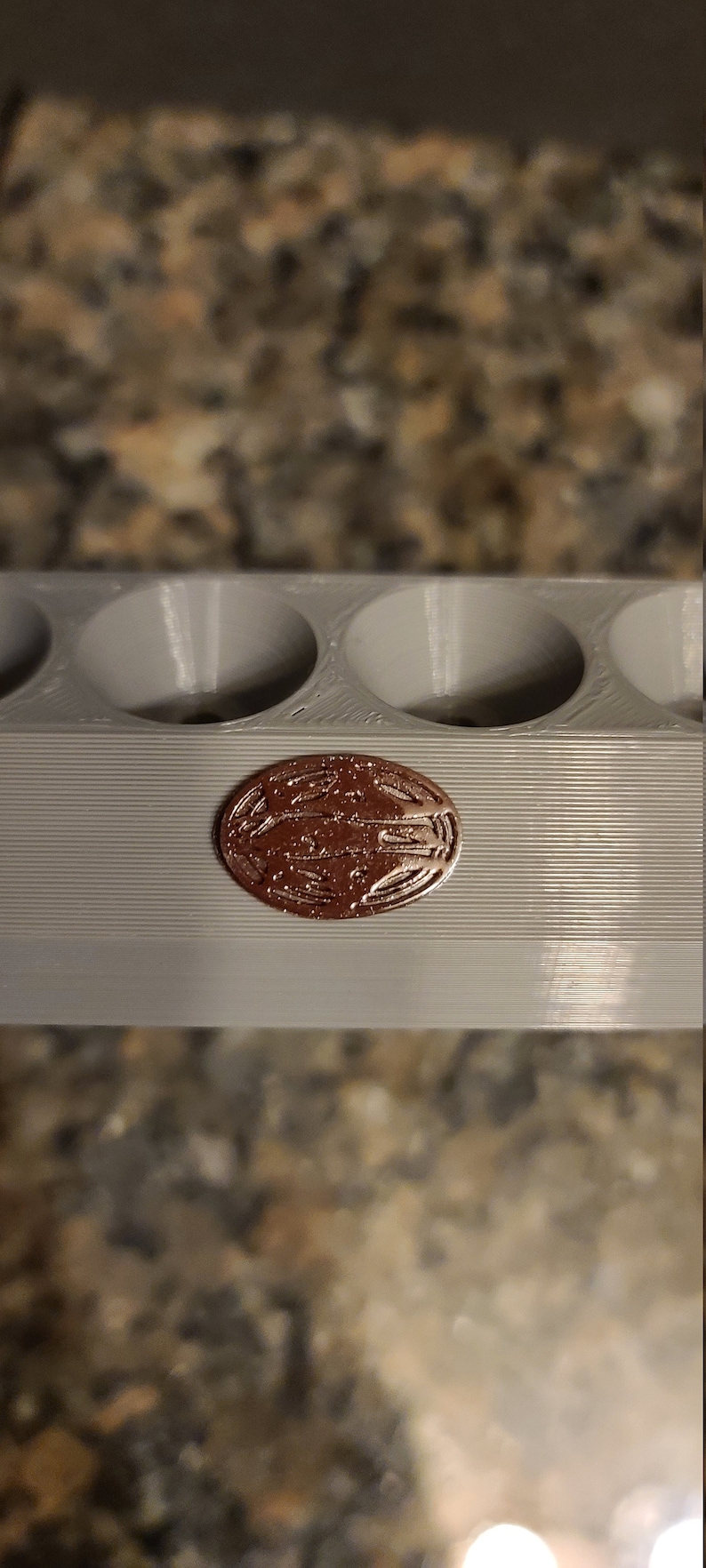 May include: A close-up of a grey plastic mould with six circular indentations. A copper-coloured coin with a design is sitting in one of the indentations.