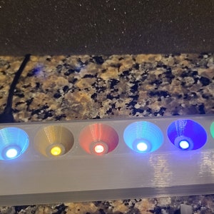 May include: A grey 3D printed stand with six small, coloured lights. The lights are in a row and are lit up in different colours, including blue, green, yellow, and red. The stand has two arms on either side, each with a circular opening at the top.