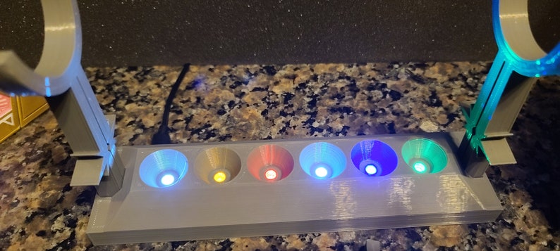 May include: A grey 3D printed stand with six small, coloured lights. The lights are in a row and are lit up in different colours, including blue, green, yellow, and red. The stand has two arms on either side, each with a circular opening at the top.