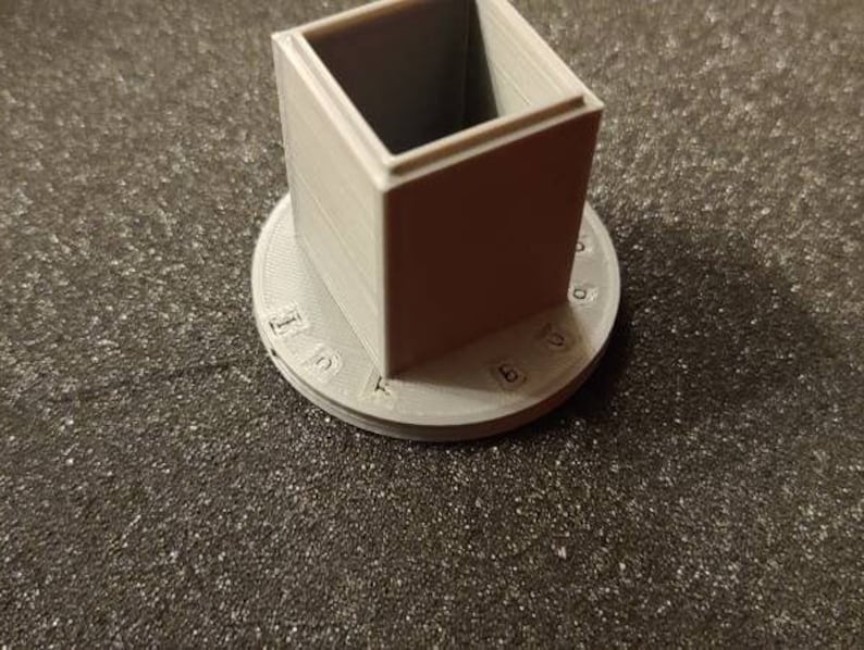 Pu&ograve; includere: Un cubo bianco stampato in 3D con una base circolare. La base ha i numeri da 1 a 10 stampati attorno al bordo.
