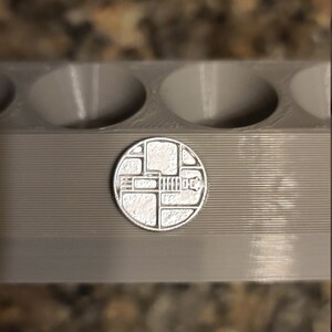 May include: A silver-coloured metal mould with four round cavities. The mould is sitting on a brown surface. A small, silver-coloured metal disc with a geometric design is in front of the mould.