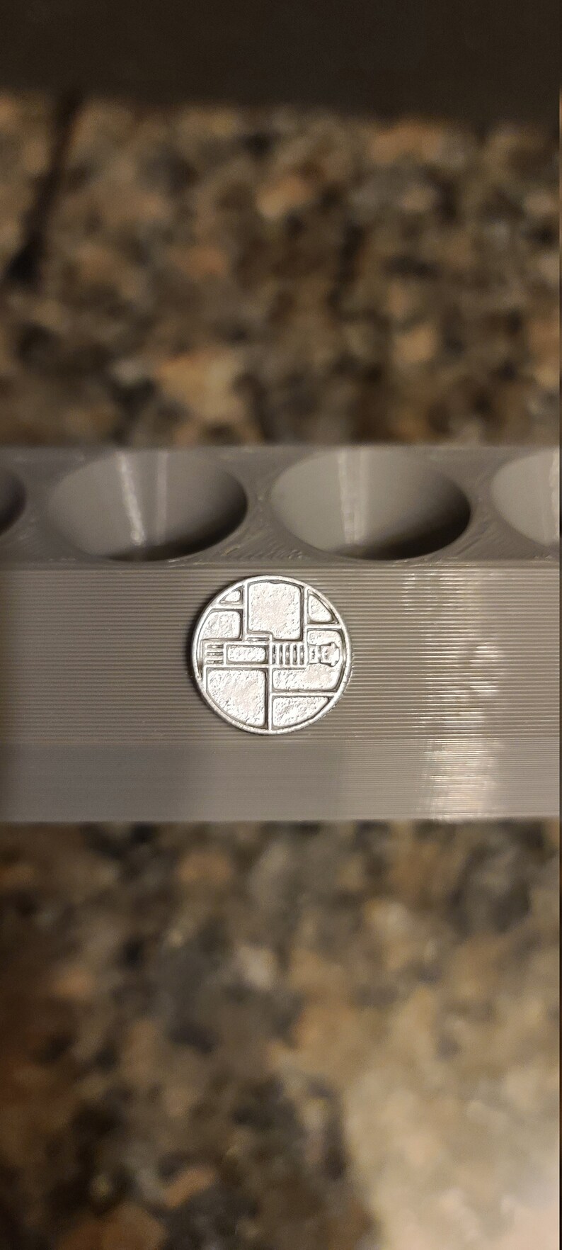 May include: A silver-coloured metal mould with four round cavities. The mould is sitting on a brown surface. A small, silver-coloured metal disc with a geometric design is in front of the mould.