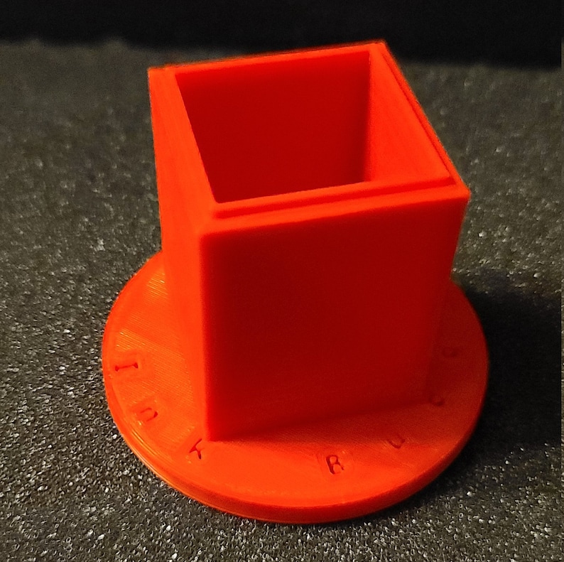 Pu&ograve; includere: Un calamaio di plastica arancione brillante con un'apertura quadrata e una base rotonda. La base ha le parole "INK" e "RULER" stampate su di essa.