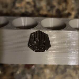 May include: A close-up of a silver-coloured metal pin with a geometric design. The pin is sitting on a grey plastic surface.