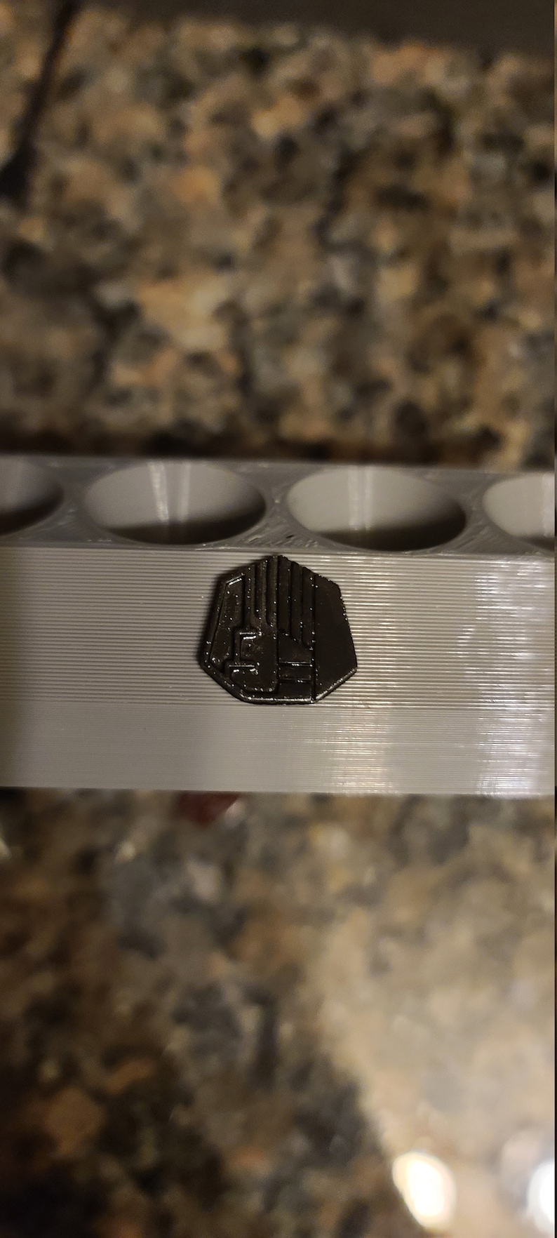 May include: A close-up of a silver-coloured metal pin with a geometric design. The pin is sitting on a grey plastic surface.