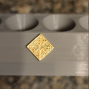 May include: A gold-coloured, diamond-shaped decorative piece with an intricate geometric pattern. The piece is sitting on a grey surface.