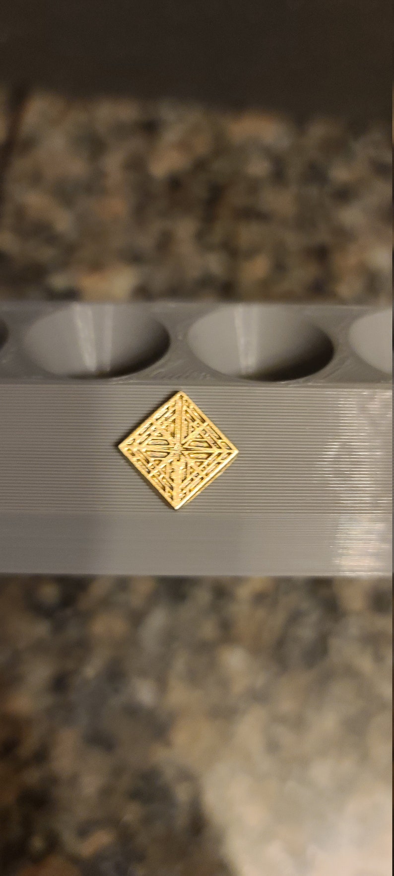 May include: A gold-coloured, diamond-shaped decorative piece with an intricate geometric pattern. The piece is sitting on a grey surface.