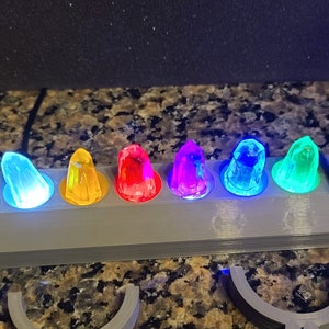 May include: A grey plastic stand with seven colourful, glowing, crystal-like objects. The stand has a raised back and a flat front with a groove for each object. The objects are arranged in a row from left to right: blue, yellow, red, purple, light blue, green, and turquoise.