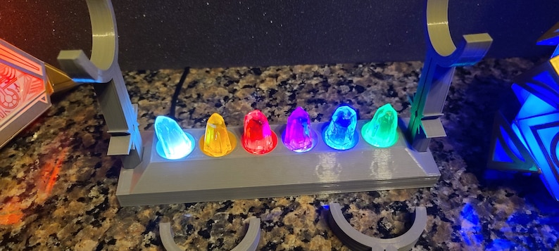 May include: A grey plastic stand with seven colourful, glowing, crystal-like objects. The stand has a raised back and a flat front with a groove for each object. The objects are arranged in a row from left to right: blue, yellow, red, purple, light blue, green, and turquoise.
