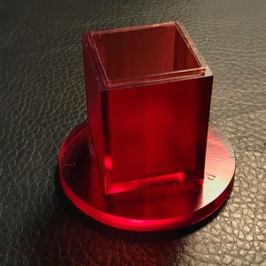 Pu&ograve; includere: Un cubo di acrilico rosso con una base quadrata si trova su una base circolare in acrilico rosso.