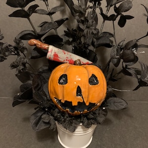 May include: A ceramic pumpkin with a carved face and a bloody knife sticking out of the top. The pumpkin is sitting in a white bucket surrounded by black flowers.