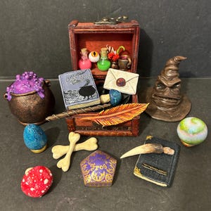 May include: A collection of miniature wizarding items, including a book titled "Advanced Potion Making", a Sorting Hat, a quill, a cauldron, a bone, a potion bottle, a crystal ball, a mushroom, and a Hogwarts house crest.