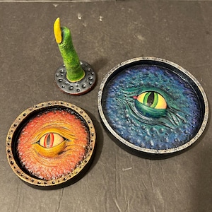 May include: Three handmade, fantasy-themed decorative items. One is a green tentacle with a yellow tip, mounted on a gray base. Two round trays feature detailed eye designs, one with a red and orange background, the other with a blue and green background.