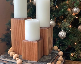 May include: Three white pillar candles of varying sizes are displayed on a wooden candle holder. The candle holder is made of natural wood with a rustic finish. The candles are arranged in a staggered pattern, with the largest candle in the back and the smallest candle in the front. The candle holder is sitting on a stack of books, and a wooden bead garland is draped around the books.