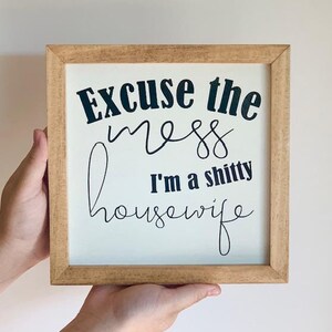 May include: A wooden sign with the text "Excuse the mess I'm a shitty housewife" in black lettering on a white background.
