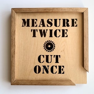 May include: A wooden sign with the text "MEASURE TWICE CUT ONCE" in black lettering. A stylized saw blade icon is between the words "TWICE" and "CUT".