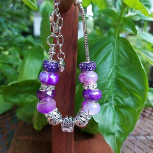 May include: A purple beaded bracelet with silver accents. The bracelet features various shades of purple beads, including some with crystal embellishments. A silver paw print charm is visible. The bracelet is displayed on a wooden stand against a backdrop of green foliage.