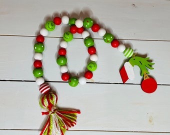 Peut inclure: Une guirlande de Noël verte et rouge avec un motif sur le thème du Grinch. La guirlande est faite de perles en bois avec des visages peints et un pompon vert et blanc.