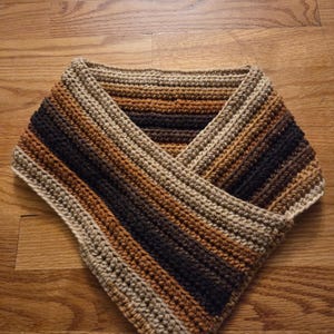 May include: A handmade crochet cowl in a V-neck style. The cowl features horizontal stripes in shades of brown, tan, and black. The yarn appears to be a soft, textured material. The cowl is designed to be worn around the neck.