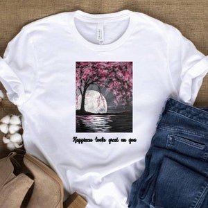 May include: White t-shirt with a rectangular graphic featuring a tree with pink blossoms, a full moon, and water reflection. The text "Happiness looks great on you" is below the graphic. The shirt is paired with blue jeans and beige shoes.