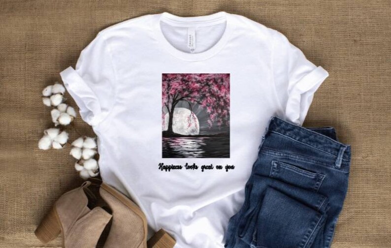 May include: White t-shirt with a rectangular graphic featuring a tree with pink blossoms, a full moon, and water reflection. The text "Happiness looks great on you" is below the graphic. The shirt is paired with blue jeans and beige shoes.